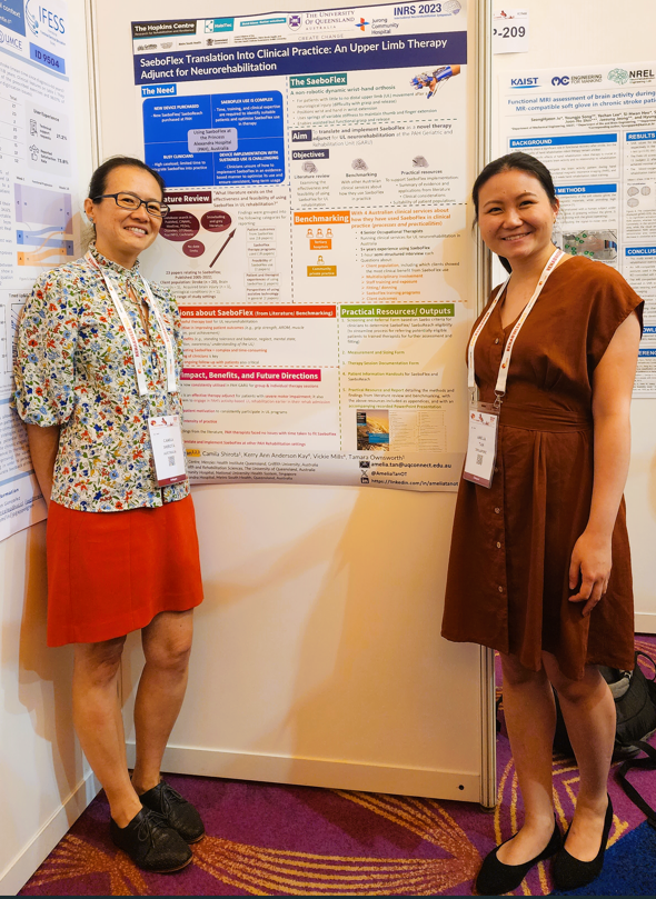 Camila Shirota, a woman with dark hair and glasses stands in the corner of the poster presentation area. Amelia Tan, a woman with dark hair and wearing a brown dress stands to the right of her poster entitled "SaeboFlex Translation into Clinical Practice: An Upper Limb Therapy Adjunct for Neurorehabilitation"