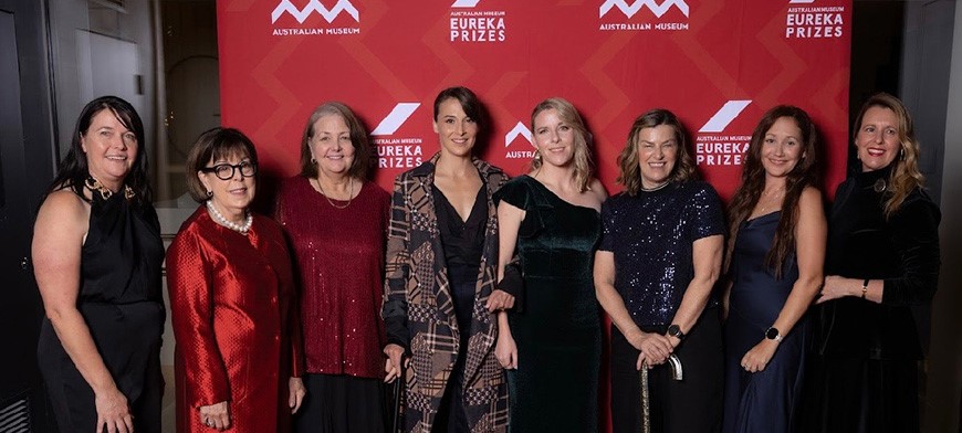 Kelsey Chapman, her PhD supervisors and other Dignity Project team members standing together at the Eureka Prize gala event. Behind them is a red background wall, with white text which reads: Eureka Prize 