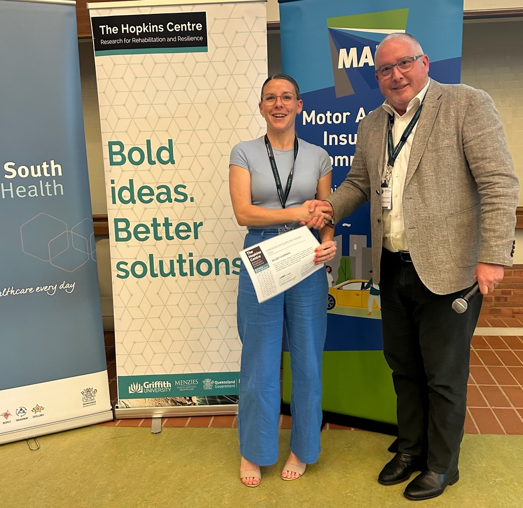A photo image of Kelsey Chapman shaking hands with Professor Tim Geraghty, in front of a Metro South Health banner and a green Bold Ideas Better Solutions Banner