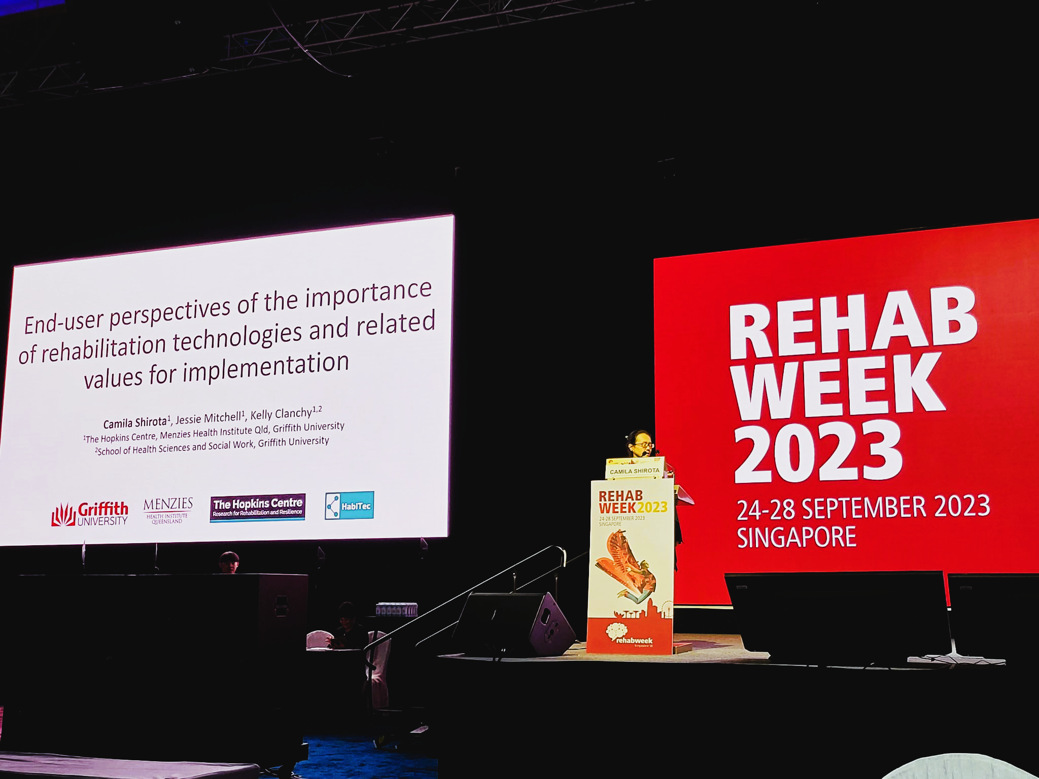Camila Shirota stands behind a lectern on a large stage. On the front of the lectern is a rectangular poster and behind her is a large billboard, both with the words “REHAB WEEK 2023” written in bold. To Camila’s right hand side is a large screen showing the title slide from her presentation “End-user Perspectives of the Importance of Rehabilitation Technologies and Related Values for Implementation”  