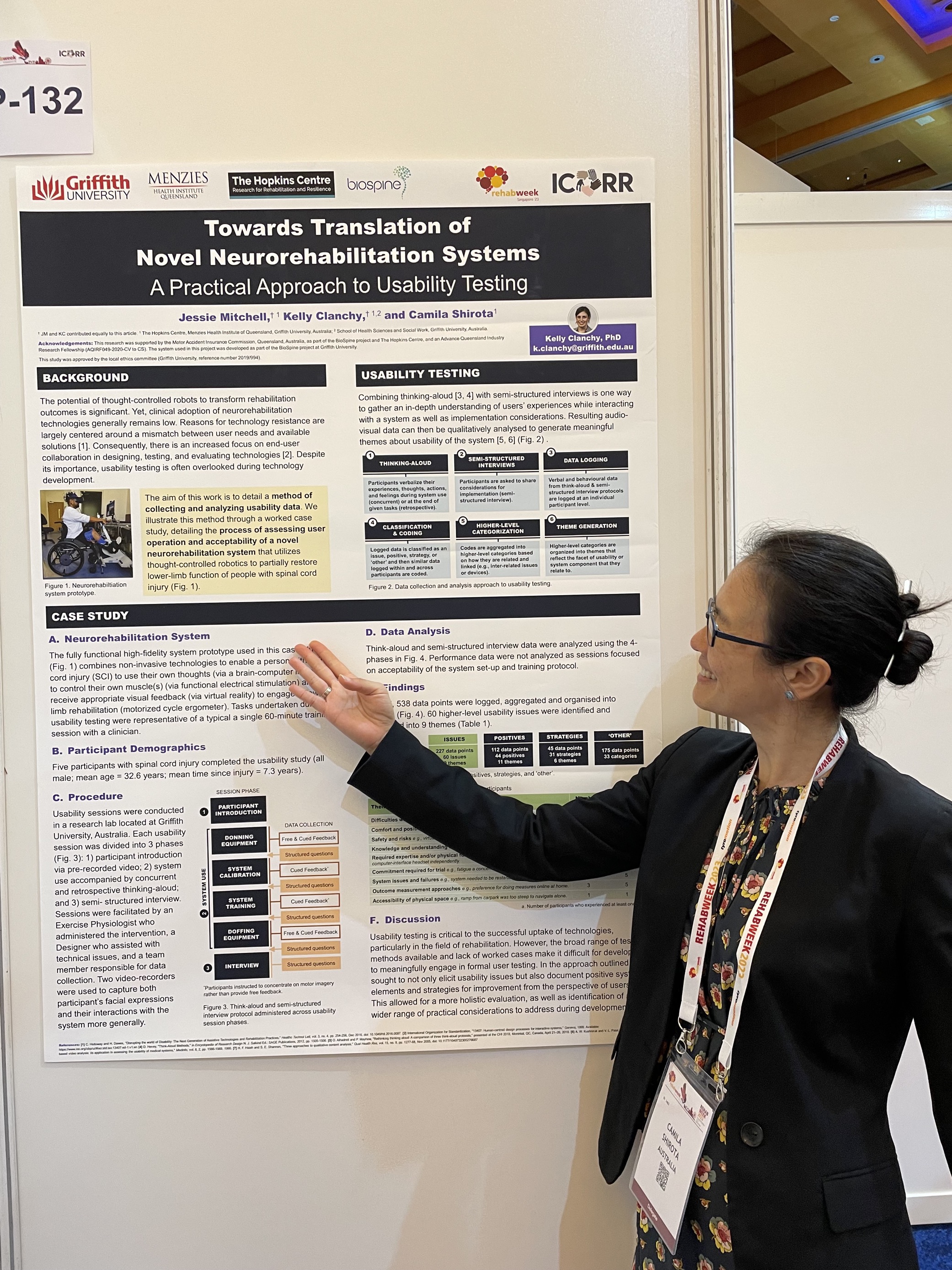 Camila Shirota, a woman with dark hair and glasses, wearing a floral dress with a black jacket faces side on to the camera, looking at the poster entitled “Towards Translation of Novel Neurorehabilitation Systems – A practical Approach to Usability Testing” 