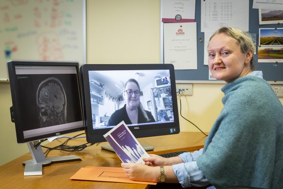 Alt text: Tamara Ownsworth - a woman with tied back, blonde hair and wearing a green jumper - sits at a desk meeting with one screen showing Julia Robertson and the other screen showing Julia's brain scan.