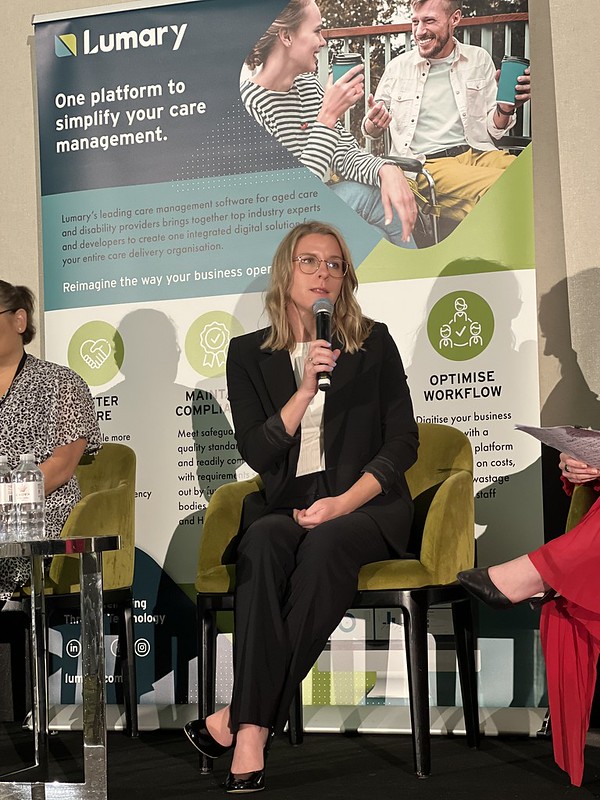 Kelsey Chapman sits on a stage in front of a large green sign, speaking into a microphone 