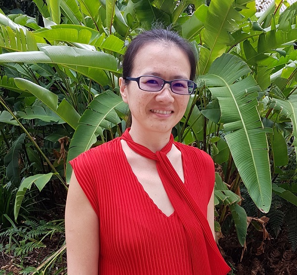 Camila Shirota, a woman with black hair and glasses, stands in front of large green leaves, wearing a red top and matching scarf