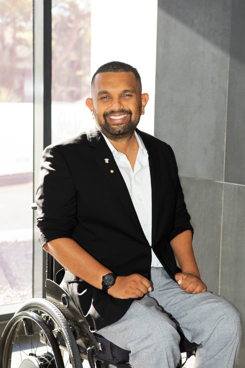 Dinesh Palipana wearing a black jacket, grey pants and a white top, sitting in his wheelchair near a large window