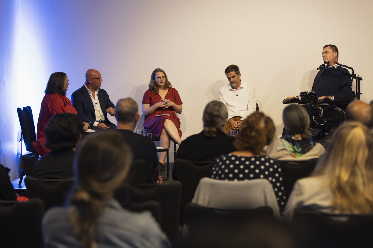 Image of THC researcher Coral Gillett and ambassador Alison McDonald sitting in behind a coffee table on stage with other panellists at the Gold Coast Open House talks series. There is a blue light in the background.