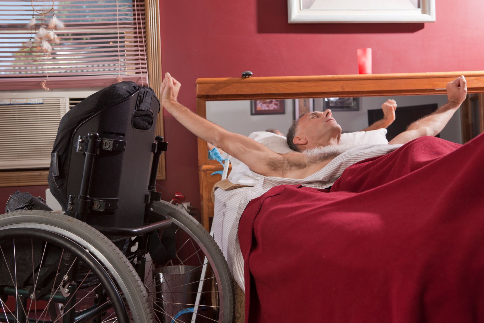 A tile with photo image at the top where a man lays in a bed covered by a red blanket, stretching his arms. A wheelchair is sitting to the left of the bed. Below the image is a white banner with turquoise text reading "2024 Seed Grant Spotlight - Seep Disturbance Following SCI"