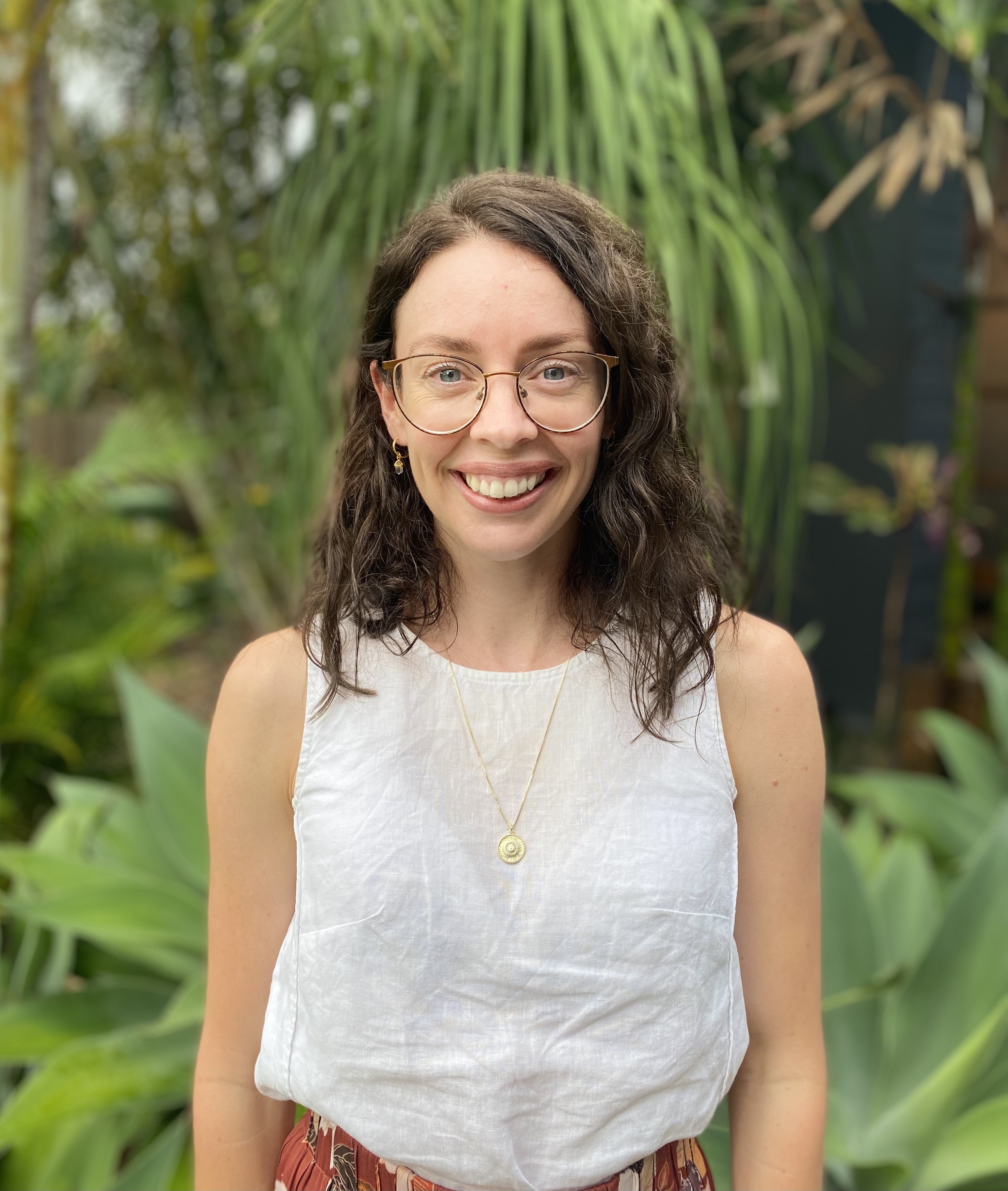 Hannah Kennedy, a woman with long brown hair, a white shirt and round glasses, stands smiling in front of a green space.