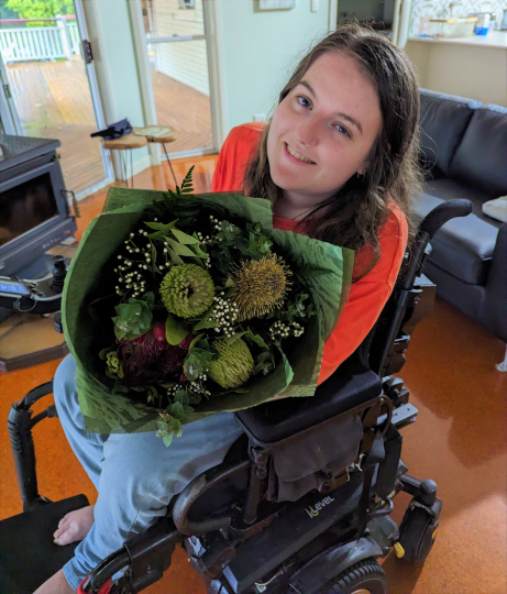 Hannah Gawne, a woman with brown hair, wearing an orange long sleeve shirt and jeans, sits in her wheelchair in a loungeroom. She is smiling at the camera and holding a large bunch of native Australian flowers. 