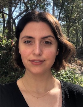 Jessie Mitchell, a woman with short brown hair, wears a black shirt and a thin gold necklace and stands in front of a natural space.