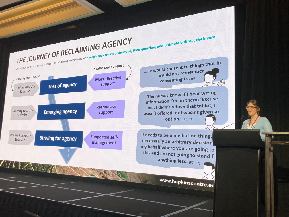 Jessie Mitchell presenting in front of an audience, at the ASSBI conference. Jessie stands behind a lecturn to the right hand side, to the left side of the picture is a large screen with slides from her presentation.