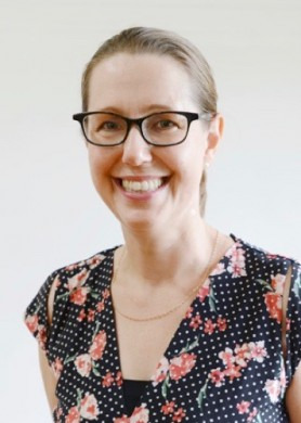 a photo image of Dr Kerrin Watter, a woman with long brown hair wearing black rimmed glasses, a black dress with white dots and flowers, and a necklace. A white wall can be seen in the background. 
