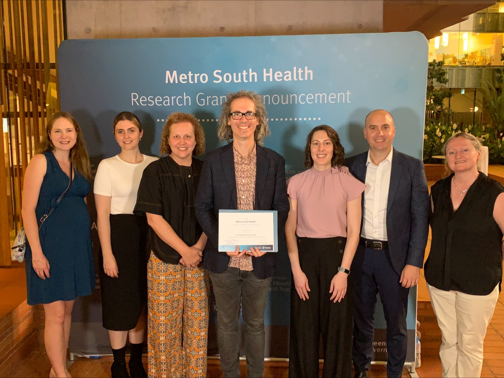A photo image of a team of researchers, smiling, in front of a Metro South Health banner.