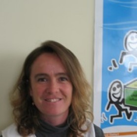 Melissa Kendall, a woman with shoulder-length, wavy, sandy blonde hair stands in front of a blue poster.