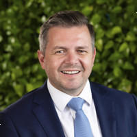 Michael Dobbie-Bridges wearing a suit and tie, sitting in front of a green bush