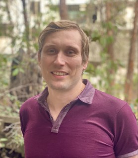 Mike Norwood, a man with short blonde hair, wearing a maroon shirt, stands in front of a natural green space.