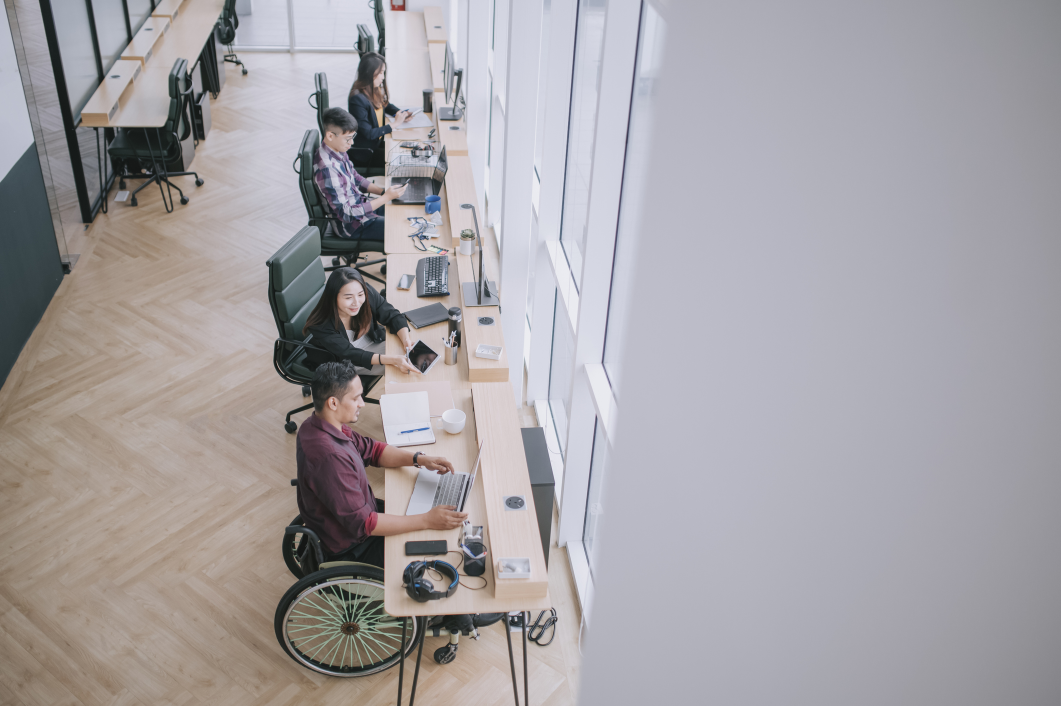 People sit along a set of windows at a shared desk, in a brightly lit room. One person sits in a wheelchair using his laptop while the others are sitting in office chairs. The man in the wheelchair and the woman next to him are speaking.. 