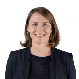 Coral Gillett, a woman with sandy blonde hair, wearing a black shirt and jacket and round earings stands in front of a white background 