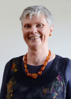 Mandy Nielsen, a woman with short grey hair, wearing a dark floral dress and orange beaded necklace.