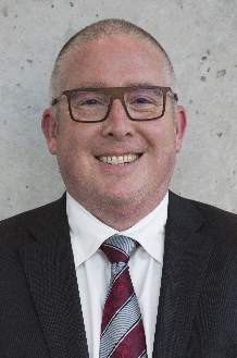 Tim Geraghty, a man with rectangular glasses, a suit jacket, fully buttoned white collared shirt and red and blue tie.